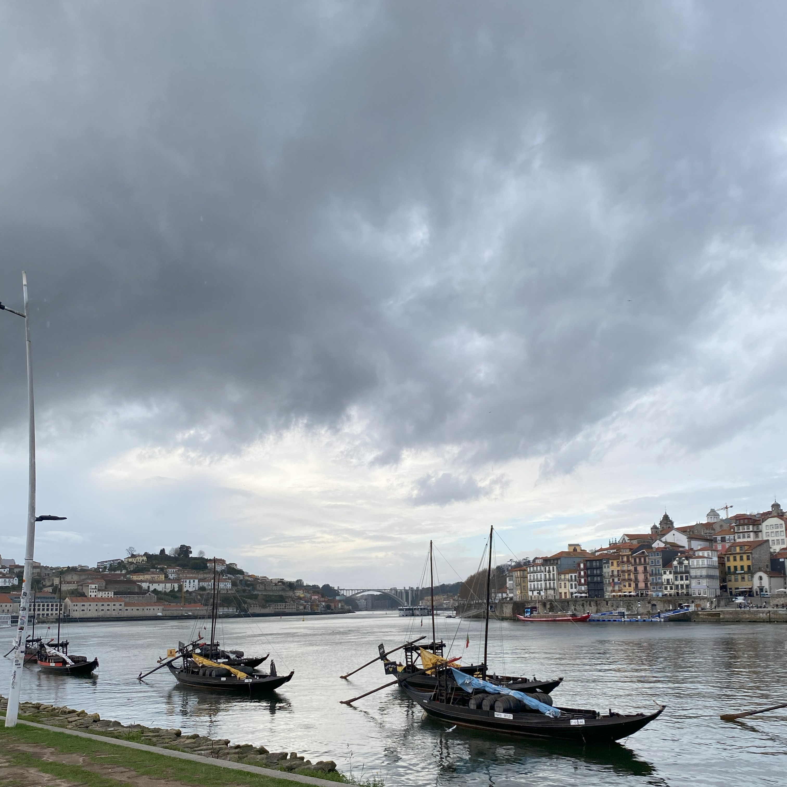 Barcos tradicionais navegando em um rio da cidade do Porto. Na margem oposta, casas coloridas e telhados de cerâmica, sob um céu nublado e cinzento.